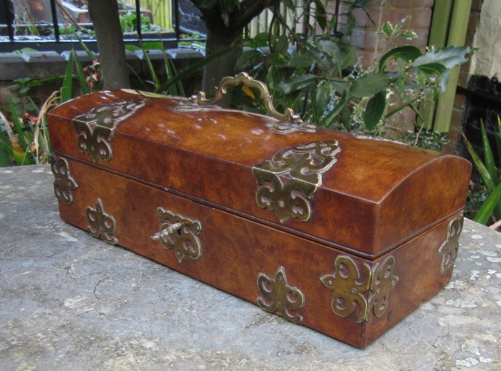 A mid-19th-century domed-top box crafted from warm brown burr walnut wood, featuring ornate pierced brass strapwork mounts, a top handle, and a key inserted into the front lock.