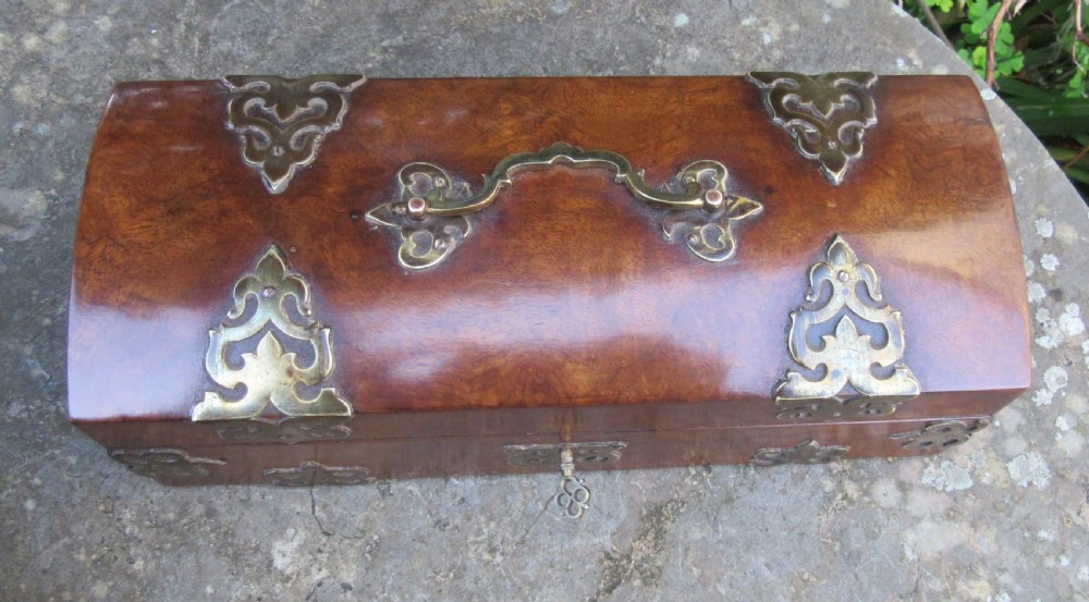 A mid-19th-century domed-top box crafted from warm brown burr walnut wood, featuring ornate pierced brass strapwork mounts, a top handle, and a key inserted into the front lock. View V