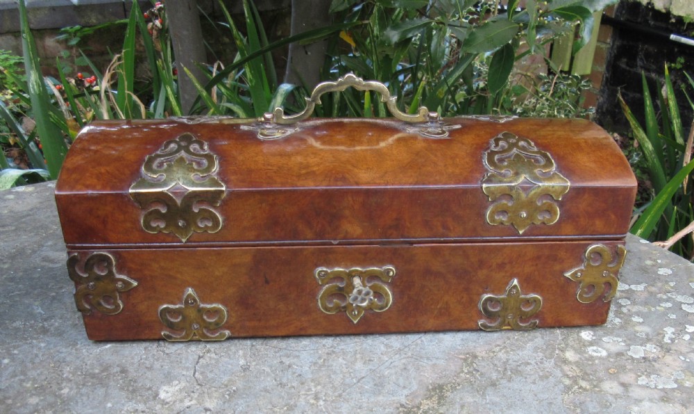 A mid-19th-century domed-top box crafted from warm brown burr walnut wood, featuring ornate pierced brass strapwork mounts, a top handle, and a key inserted into the front lock. View VI