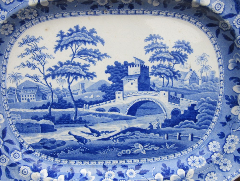 An oval blue and white earthenware serving platter with a central bridge landscape scene and a floral border. View II