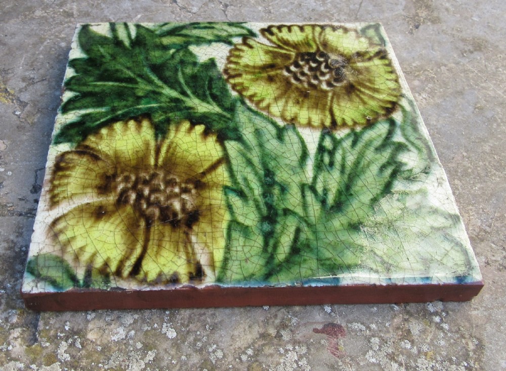 A square Arts and Crafts glazed earthenware tile featuring yellow floral motifs and green foliage with visible crazing on a terracotta base.