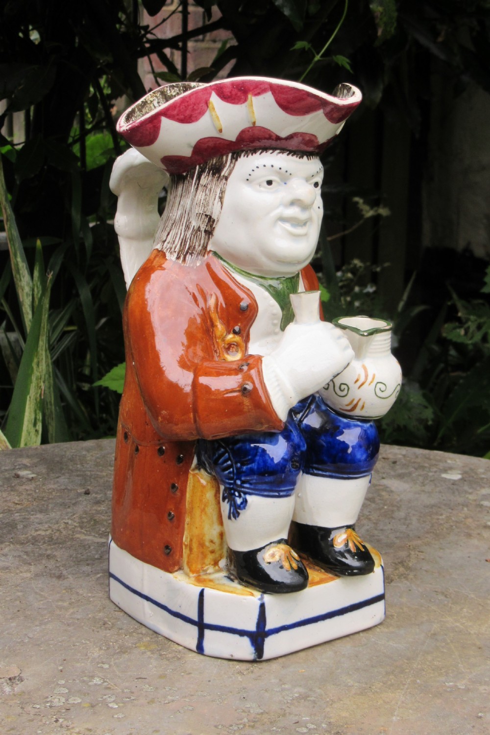 An antique early 19th-century English ceramic Toby jug depicting a seated man in a tricorn hat, brown coat, and blue breeches.