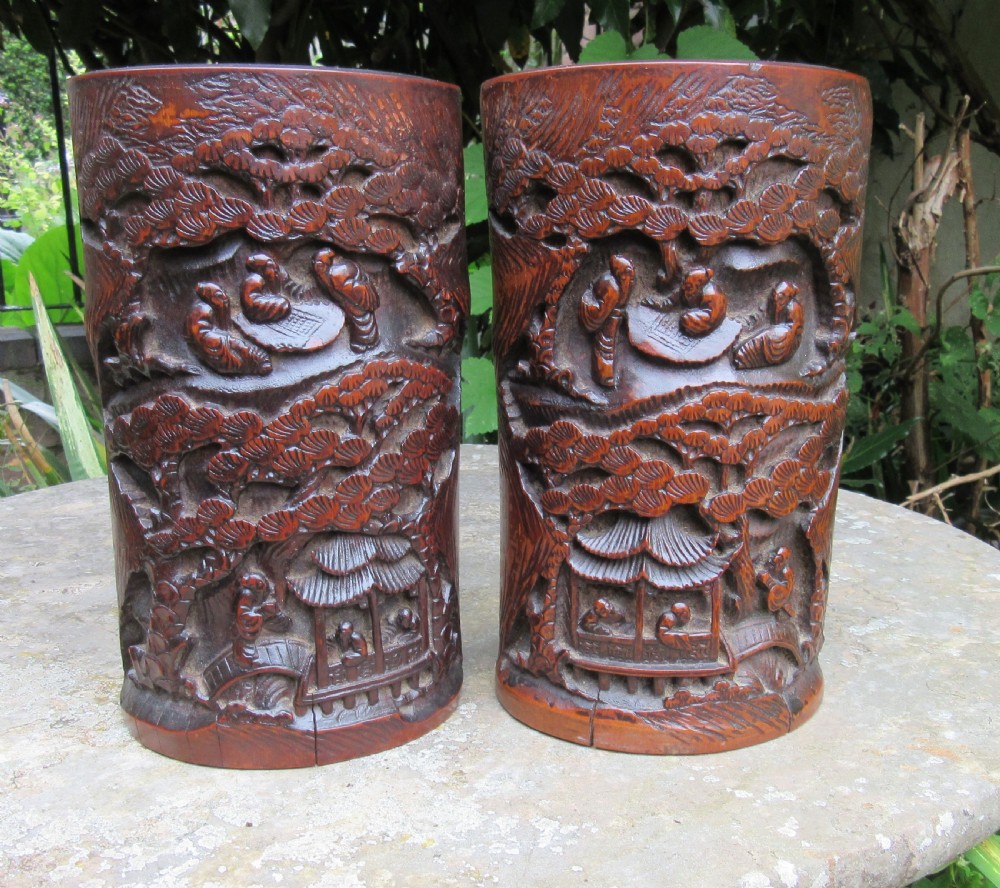 A pair of cylindrical reddish-brown carved bamboo brush pots featuring deep relief carvings of figures, pine trees, and pavilions.