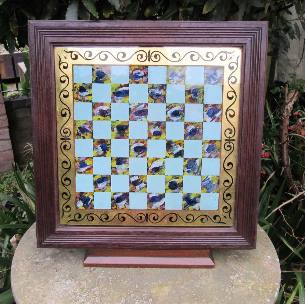 A square Victorian reverse-painted glass chess board with light blue and marbled squares, surrounded by a gilt scrollwork border and framed in dark moulded wood.