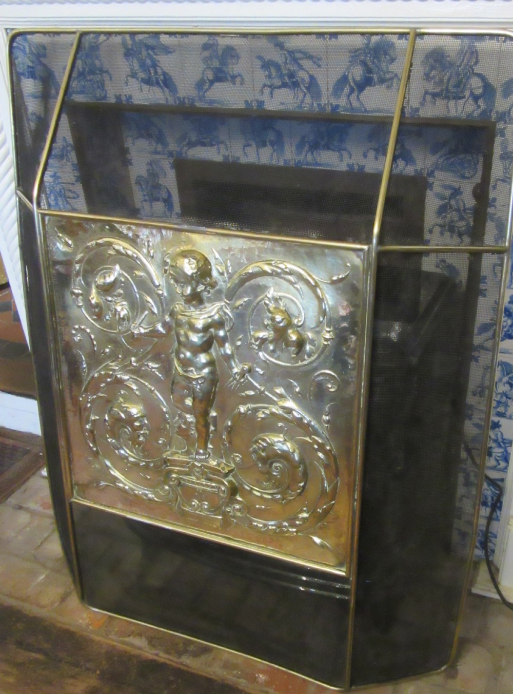 An antique embossed brass fire screen featuring a central panel with a cherub motif framed by wire mesh. View VI
