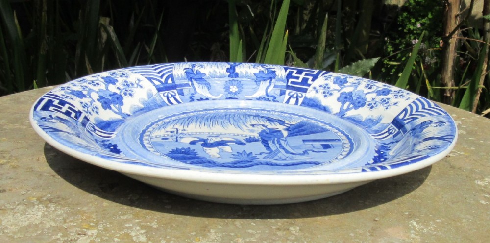 A circular antique blue and white earthenware charger decorated with a Chinoiserie landscape featuring figures and intricate geometric border patterns. View III