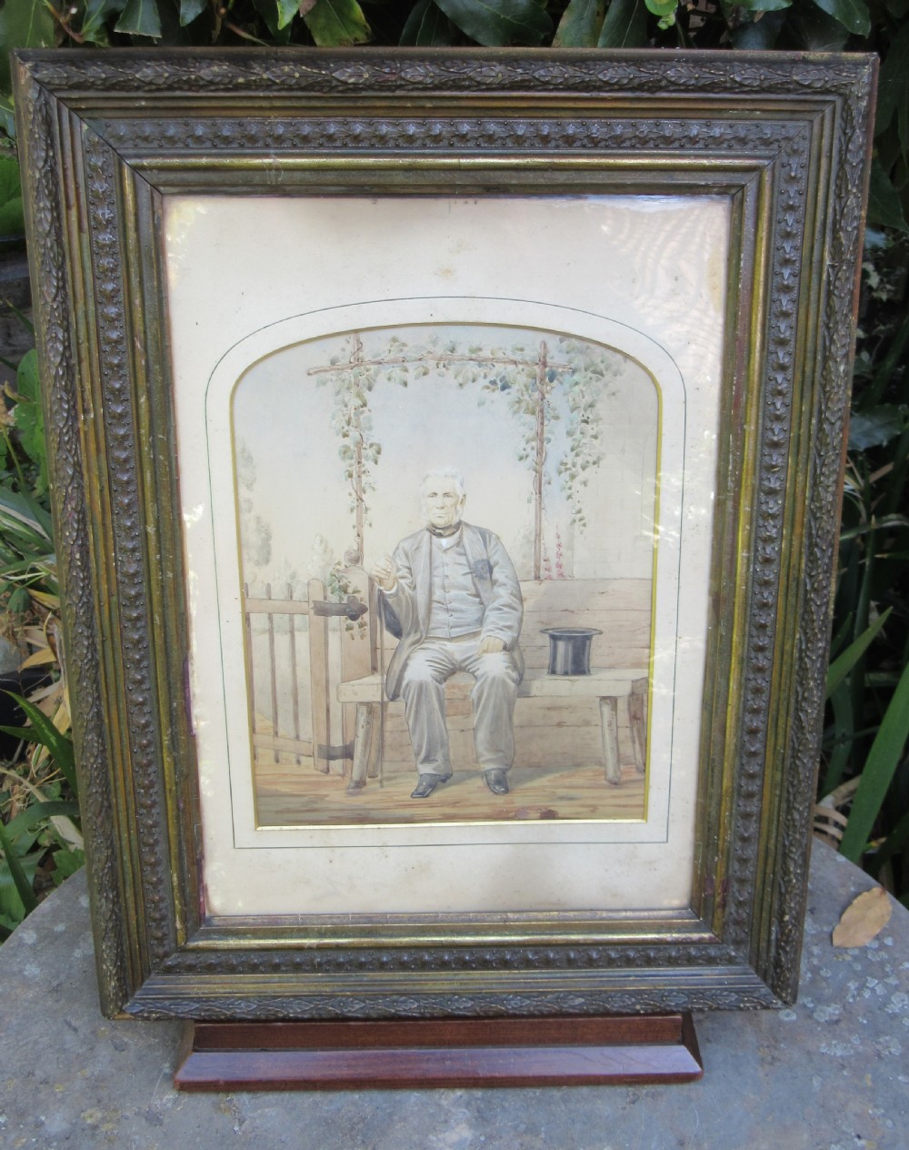 An antique primitive watercolour portrait painting depicting a seated gentleman in a garden setting within a detailed moulded frame.