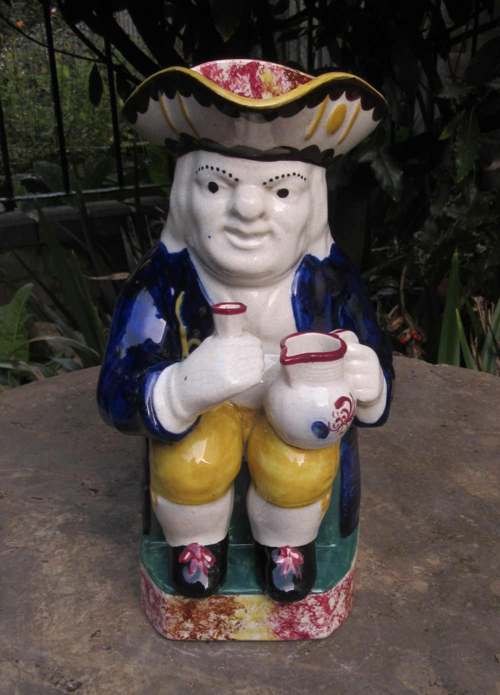 An antique English pottery Toby jug depicting a seated man in a tricorn hat, navy blue coat, and yellow breeches holding a small pitcher.