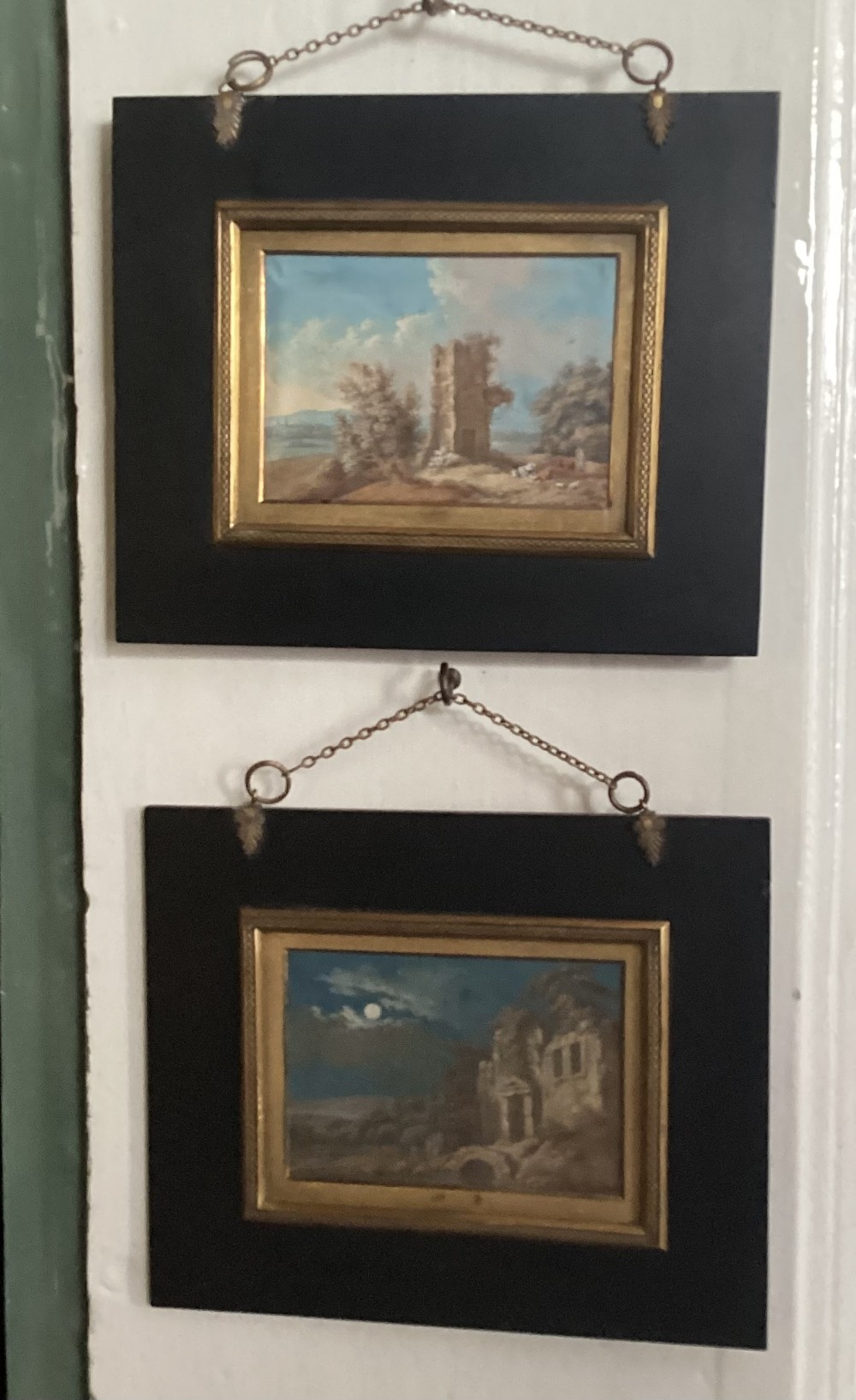 A pair of small Georgian paintings depicting architectural ruins in daylight and moonlight, set within wide ebonised frames with gilded borders and hanging chains.