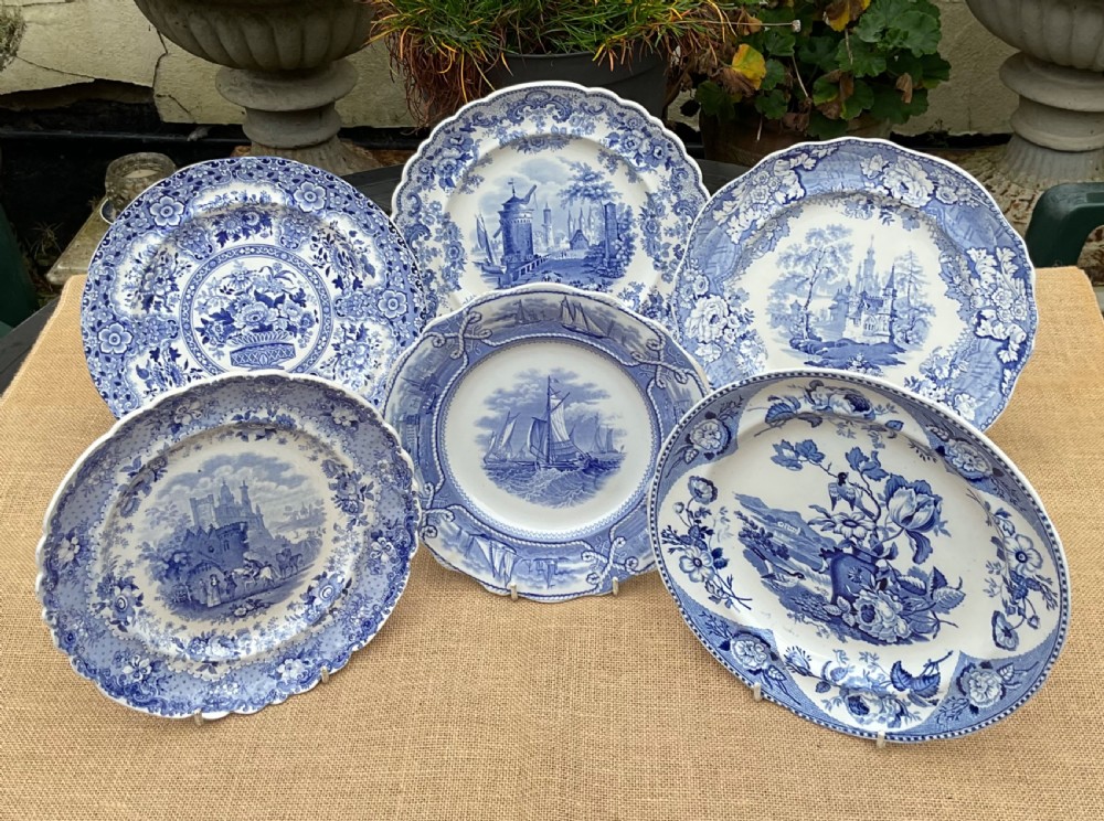 A group of six early 19th-century Staffordshire blue and white transferware ceramic plates displaying pastoral, coastal, and floral patterns with scalloped borders.