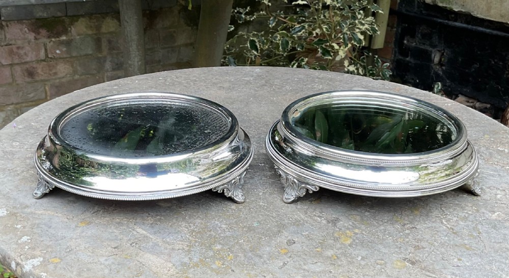 A pair of circular late Victorian silver-plated mirrored table stands featuring moulded borders, beaded rims, and ornate cast feet.