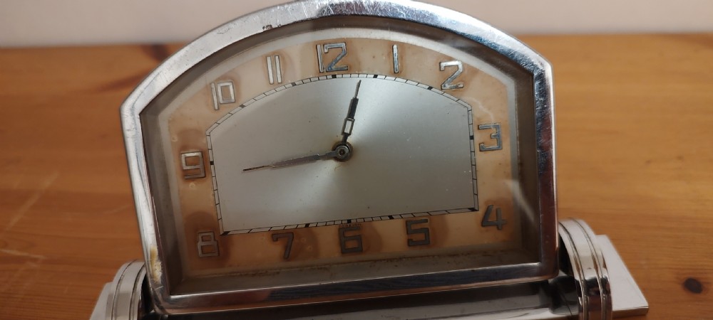 An Art Deco chrome mantel clock featuring an arched frame, stepped base, and a central dial with stylised geometric numerals. View VI