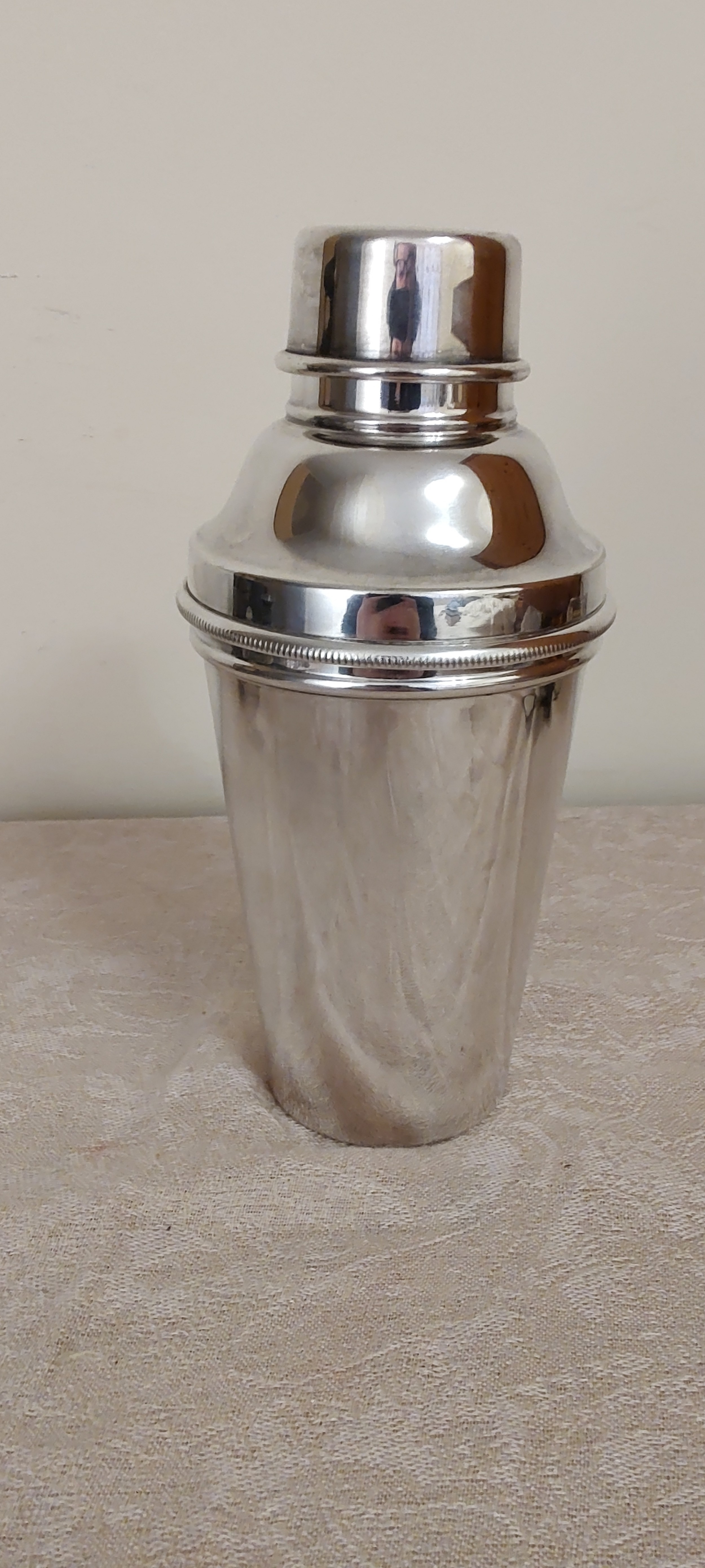 A polished silver-plated Art Deco three-piece cocktail shaker with ribbed collar details resting on a textured fabric surface.