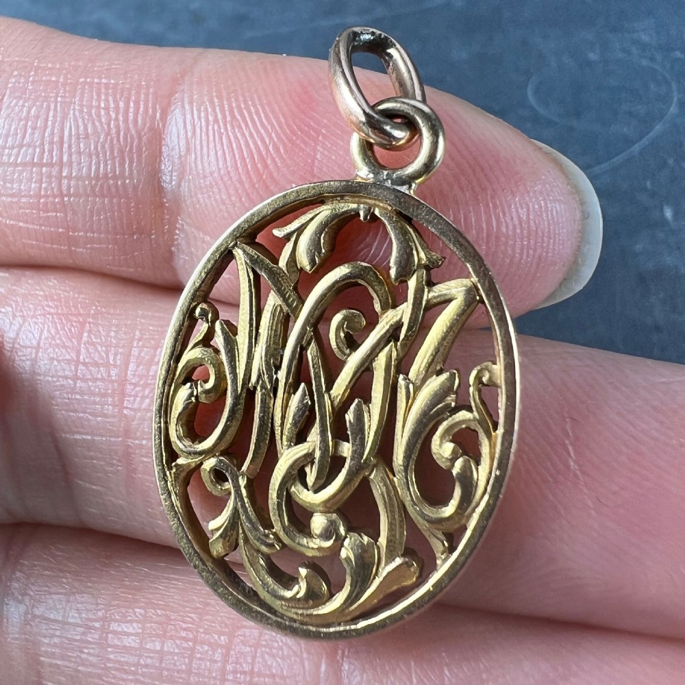An oval 18ct yellow gold pendant featuring an intricate openwork monogram within a gold frame and suspension loop. View VIII