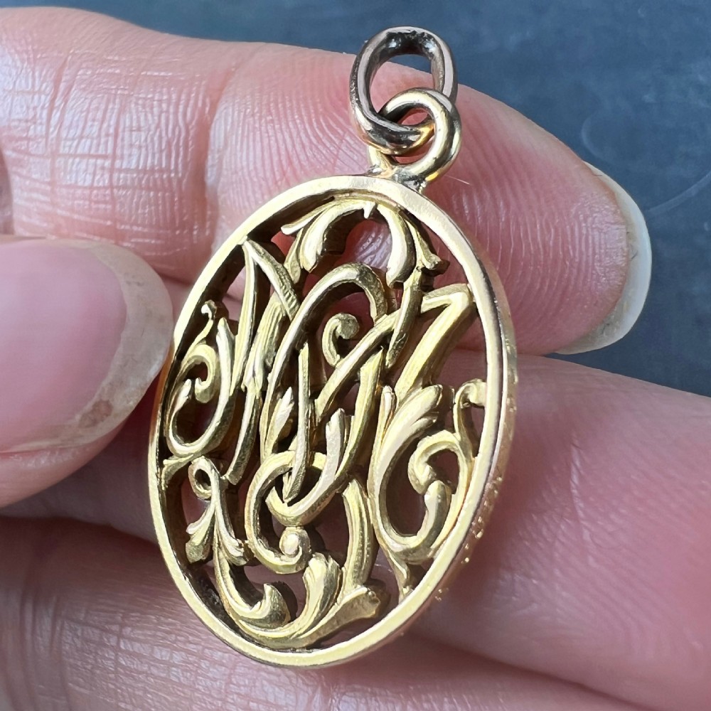 An oval 18ct yellow gold pendant featuring an intricate openwork monogram within a gold frame and suspension loop. View IX