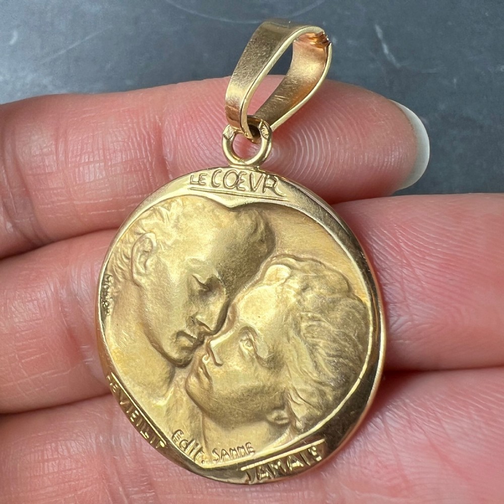 A circular 18-carat yellow gold medal pendant depicting a relief motif of two lovers kissing beneath French romantic lettering. View VI