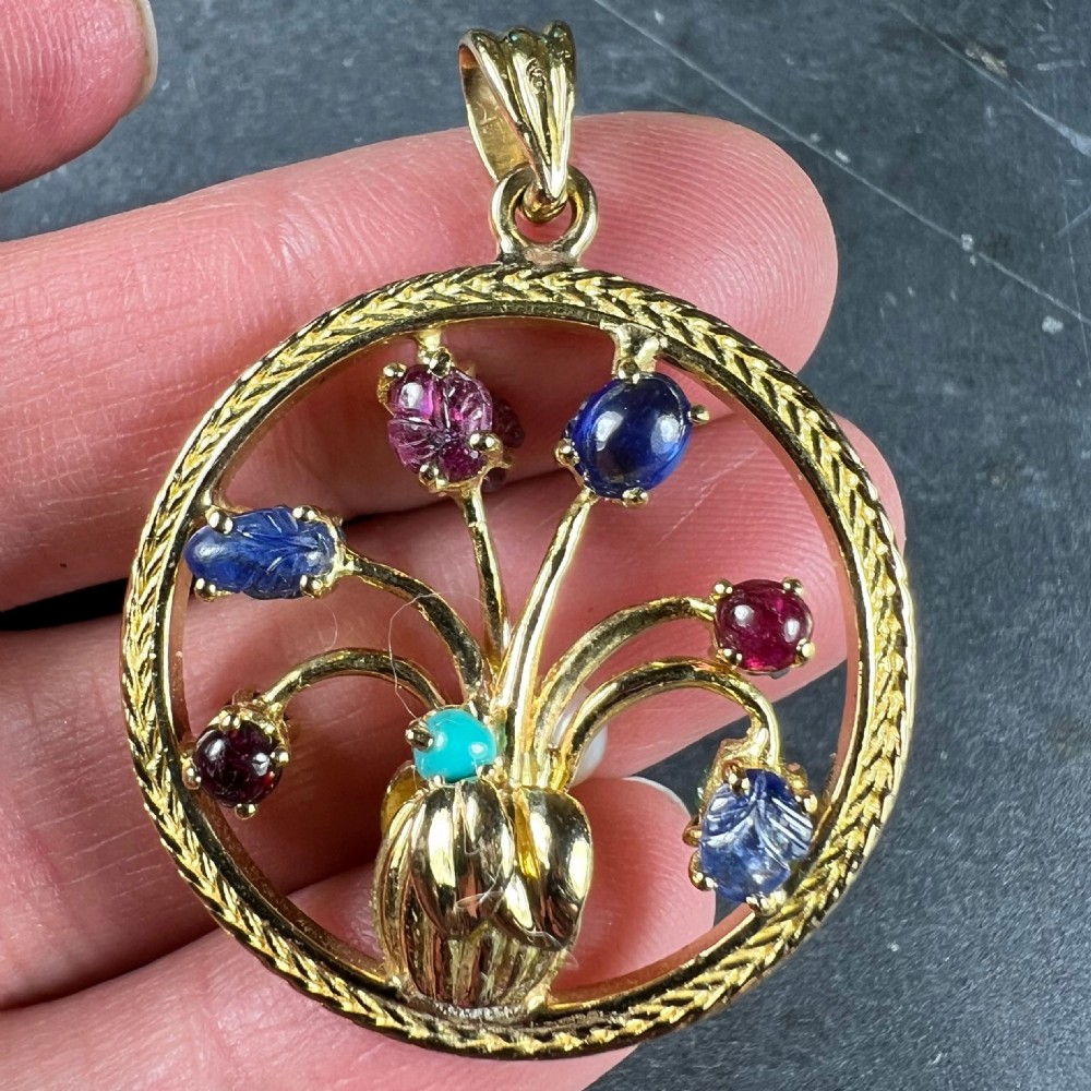 A circular 18-carat yellow gold pendant featuring an openwork flower vase design set with carved rubies, sapphires, an emerald, and a central pearl. View X