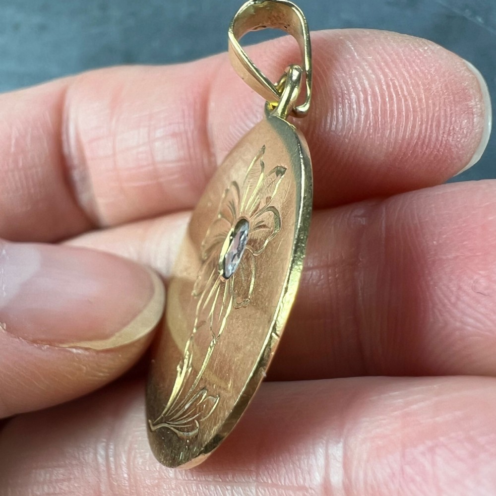 A circular 18 karat yellow gold pendant featuring an engraved daisy flower motif with a centrally set diamond and a top hanging bail. View IX
