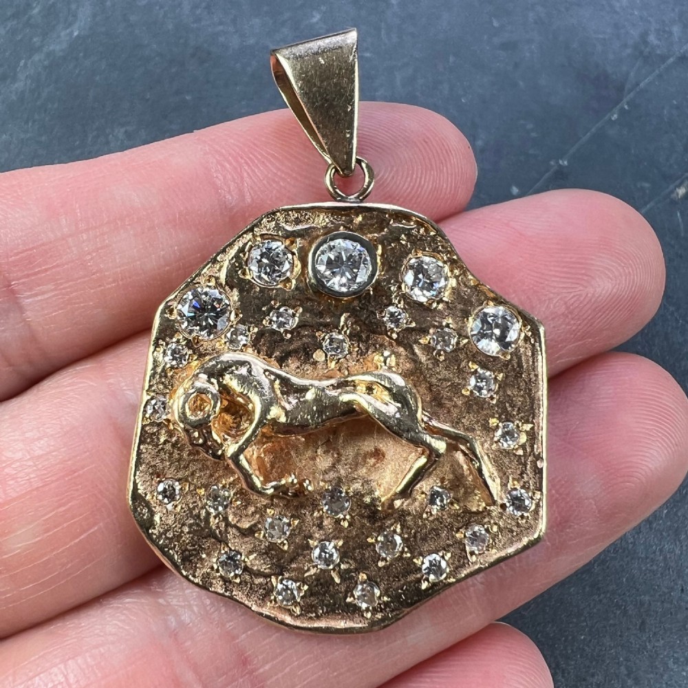A textured 14k yellow gold astrological pendant featuring an embossed horned animal motif set with small diamonds representing stars. View V