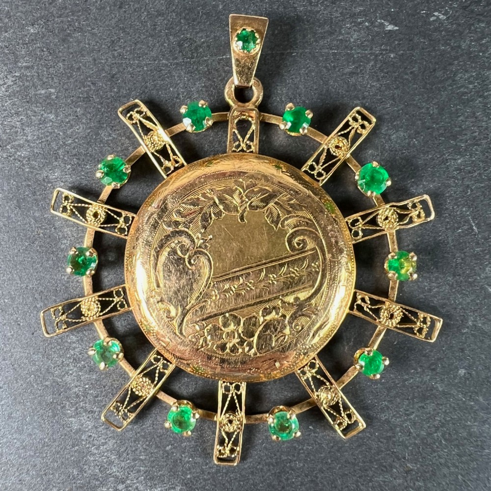 A circular 18ct yellow gold locket featuring central floral engraving and a radiating sunburst frame set with round green emeralds. View II