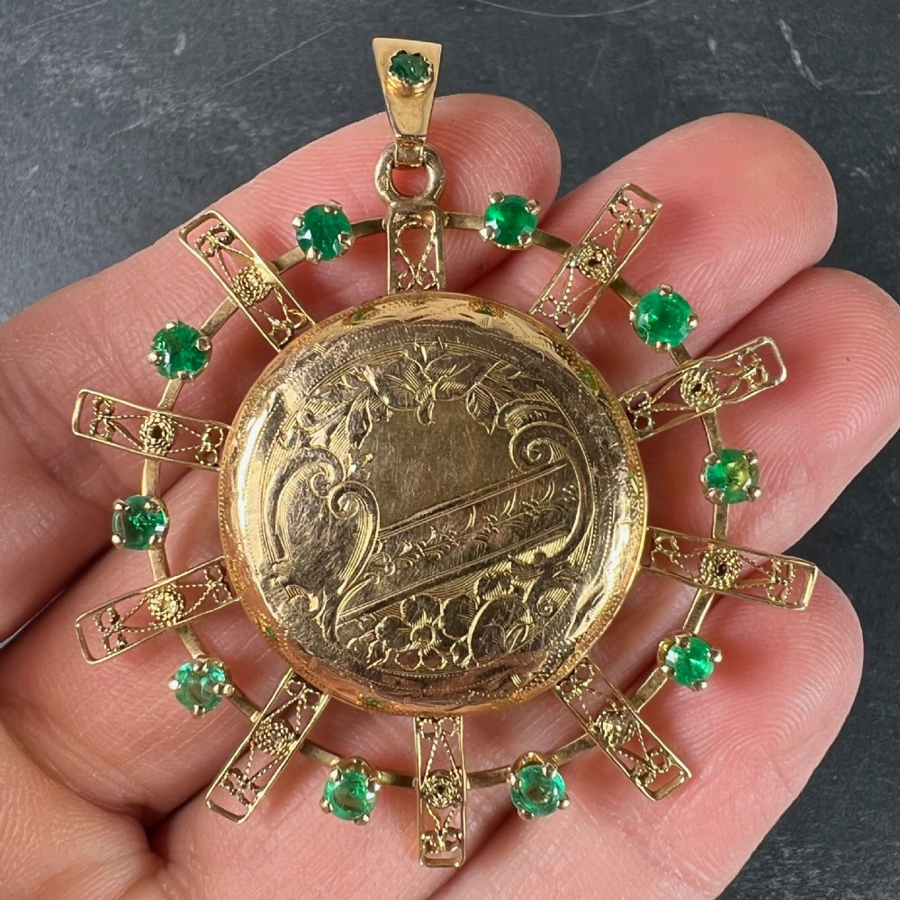 A circular 18ct yellow gold locket featuring central floral engraving and a radiating sunburst frame set with round green emeralds. View V