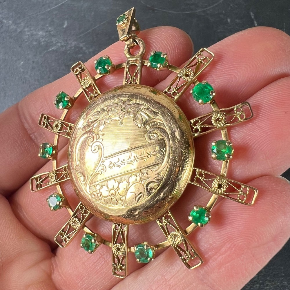 A circular 18ct yellow gold locket featuring central floral engraving and a radiating sunburst frame set with round green emeralds. View VI