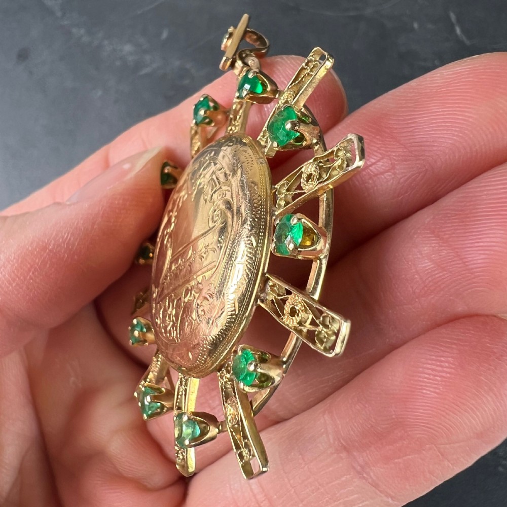 A circular 18ct yellow gold locket featuring central floral engraving and a radiating sunburst frame set with round green emeralds. View VII