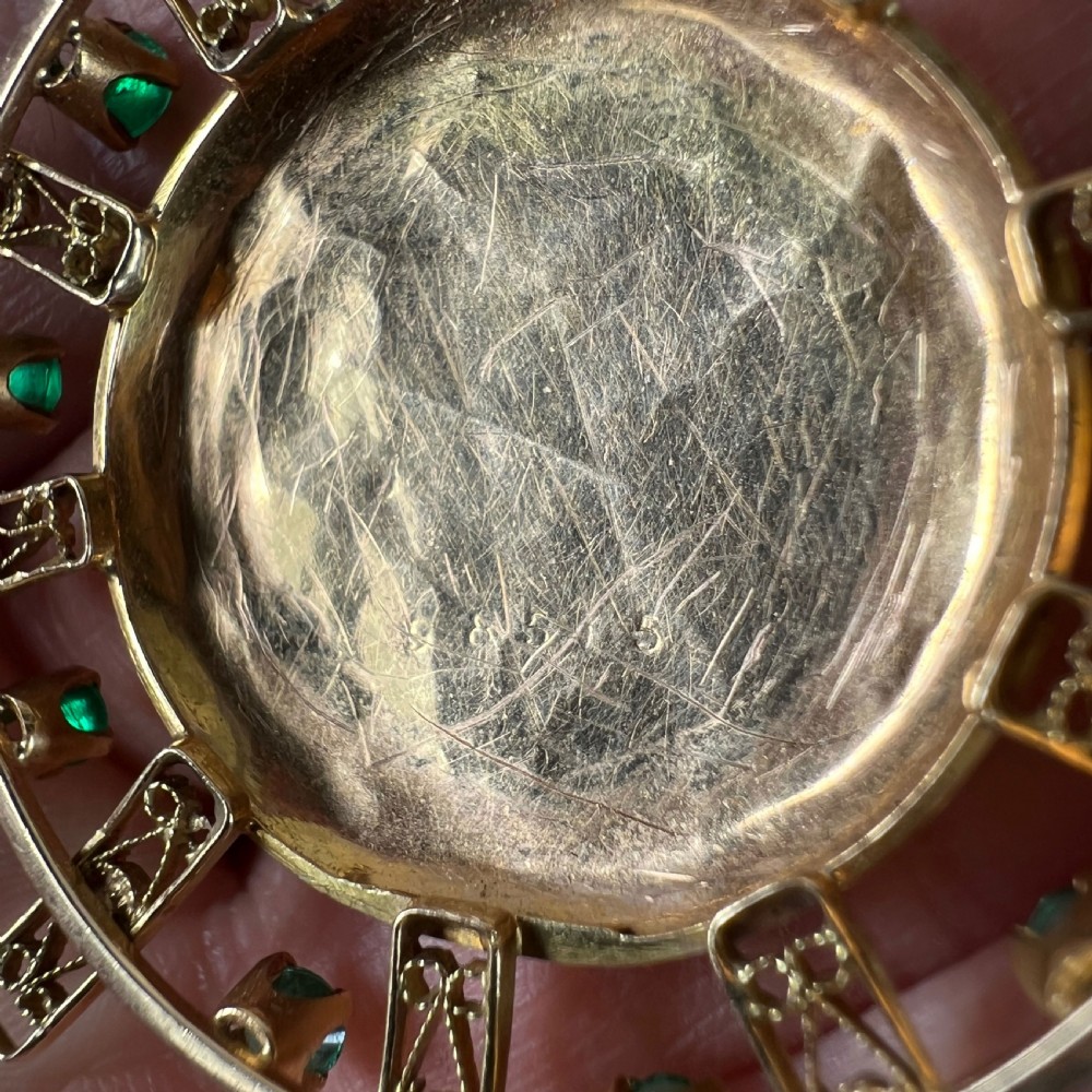 A circular 18ct yellow gold locket featuring central floral engraving and a radiating sunburst frame set with round green emeralds. View X