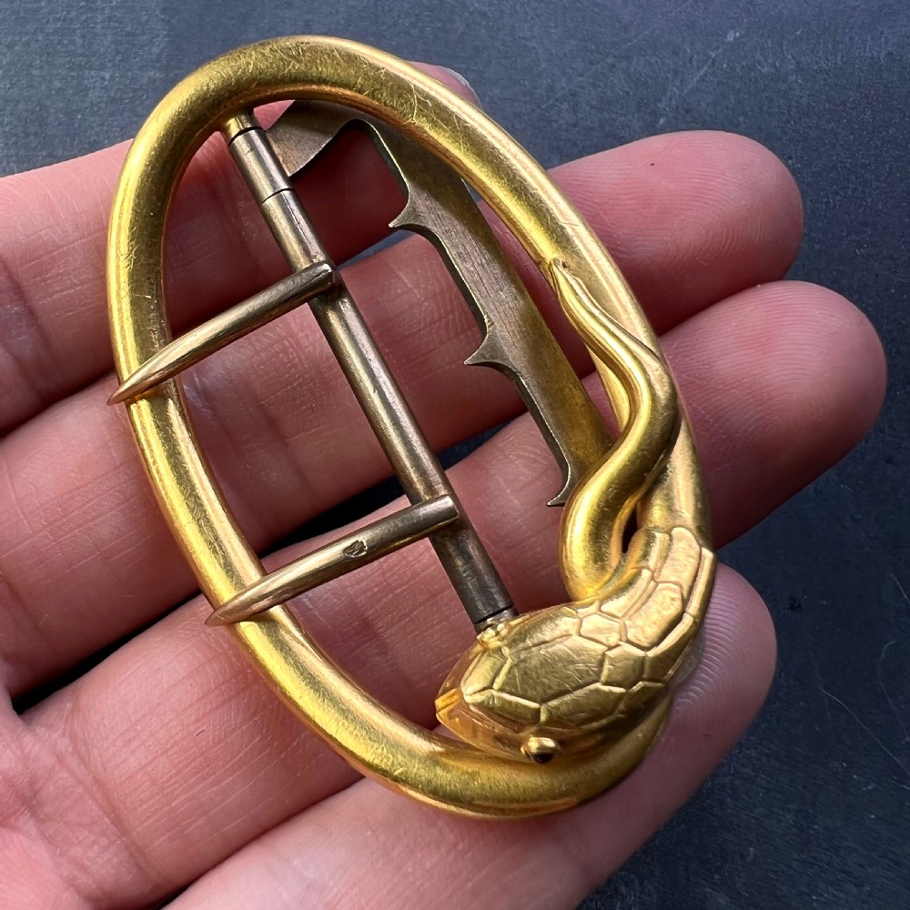 An oval 18K yellow gold belt buckle featuring a carved snake motif along the upper rim with garnet eyes. View VI