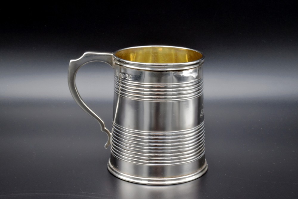 A cylindrical antique sterling silver tankard with horizontal reeded bands, a sculpted C-scroll handle, stamped hallmarks near the upper rim, and a gilded interior.