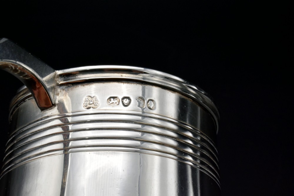A cylindrical antique sterling silver tankard with horizontal reeded bands, a sculpted C-scroll handle, stamped hallmarks near the upper rim, and a gilded interior. View V
