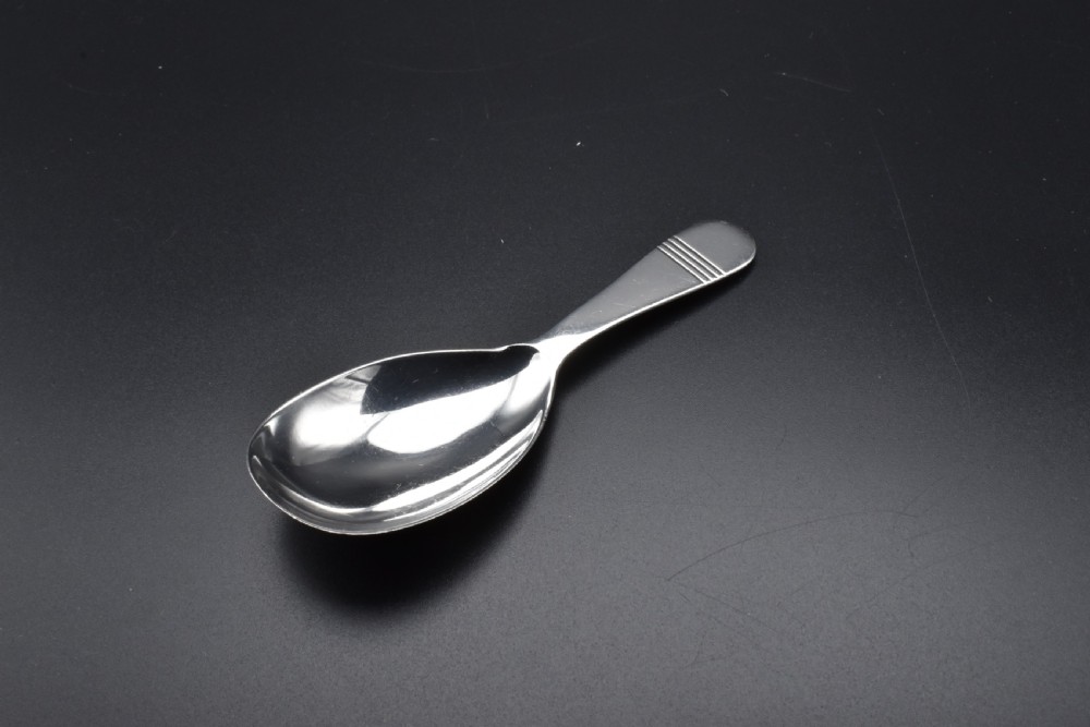 A sterling silver caddy spoon featuring a broad oval scoop bowl and a short flat handle with horizontal line accents.