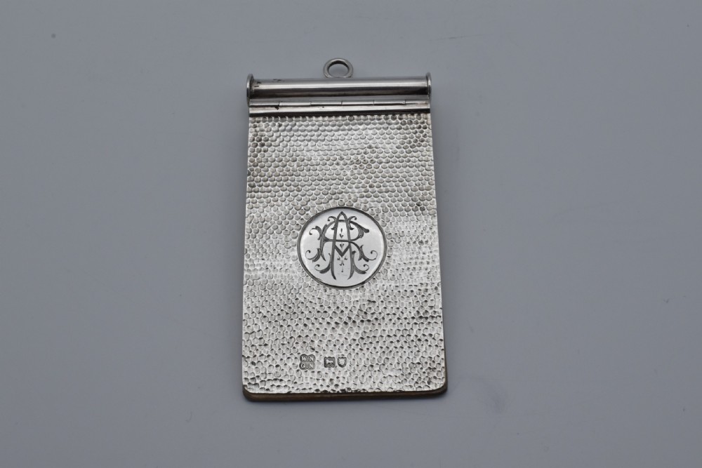 A Victorian sterling silver pocket notebook featuring a hammered finish, central monogram cartouche, top suspensory ring, and British hallmarks dated 1899.