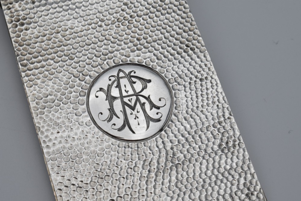 A Victorian sterling silver pocket notebook featuring a hammered finish, central monogram cartouche, top suspensory ring, and British hallmarks dated 1899. View IV