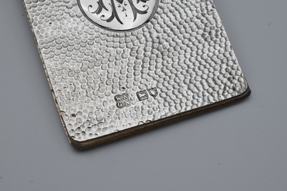 A Victorian sterling silver pocket notebook featuring a hammered finish, central monogram cartouche, top suspensory ring, and British hallmarks dated 1899. View V