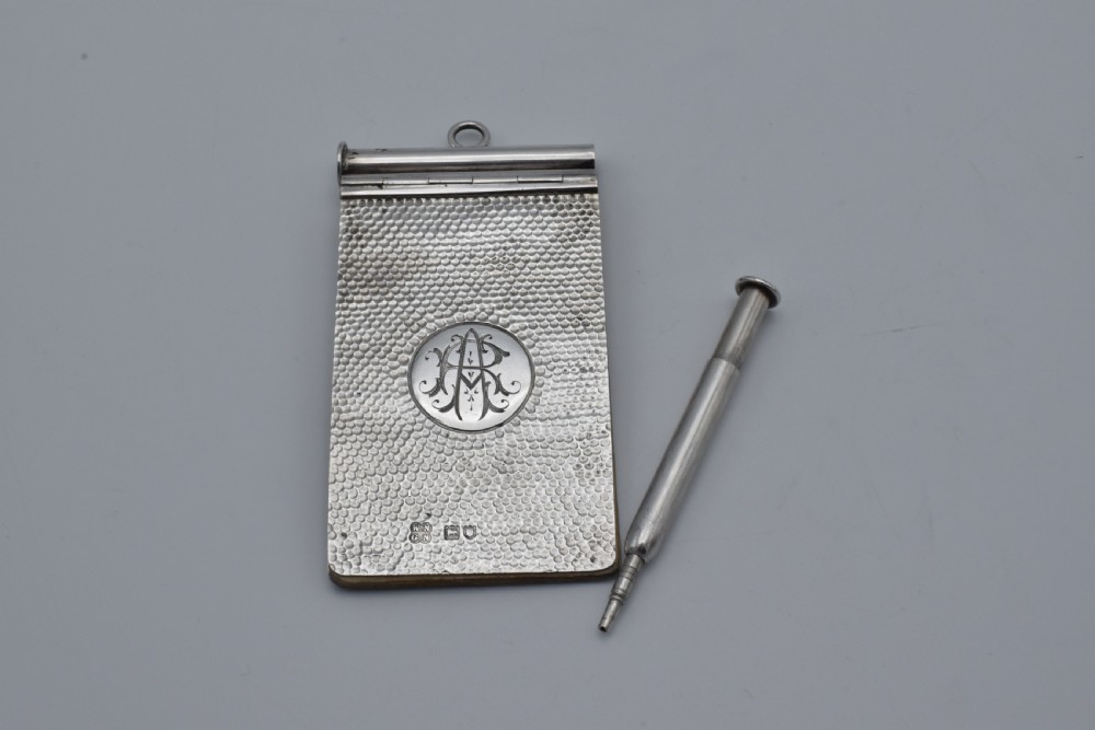A Victorian sterling silver pocket notebook featuring a hammered finish, central monogram cartouche, top suspensory ring, and British hallmarks dated 1899. View X