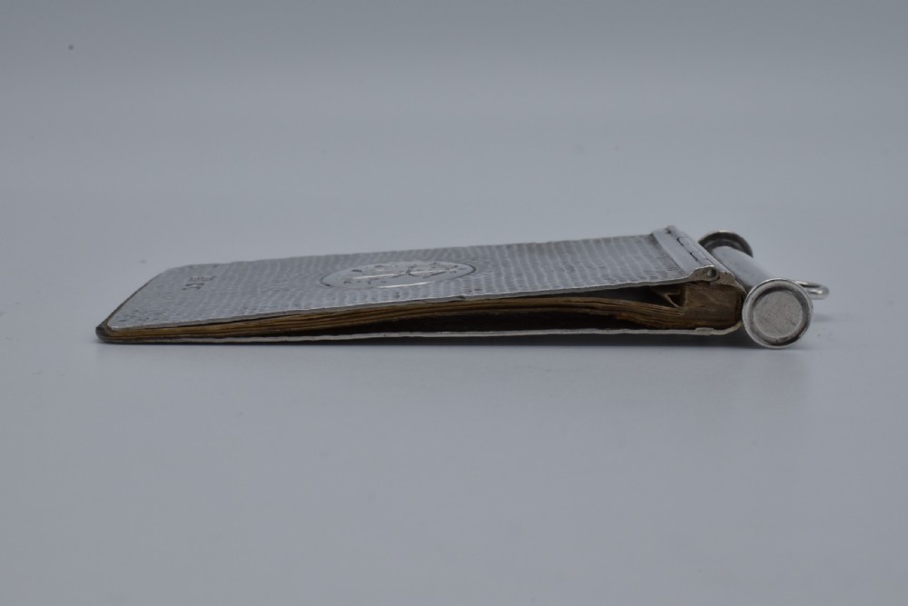 A Victorian sterling silver pocket notebook featuring a hammered finish, central monogram cartouche, top suspensory ring, and British hallmarks dated 1899. View XIII
