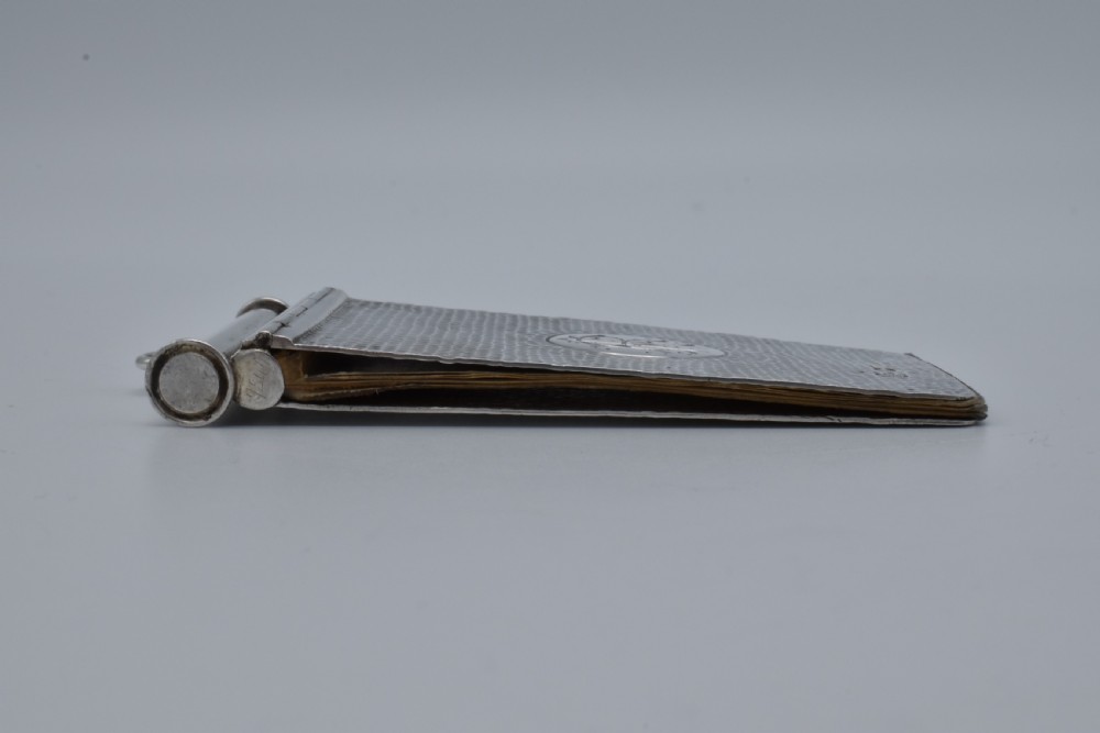A Victorian sterling silver pocket notebook featuring a hammered finish, central monogram cartouche, top suspensory ring, and British hallmarks dated 1899. View XIV