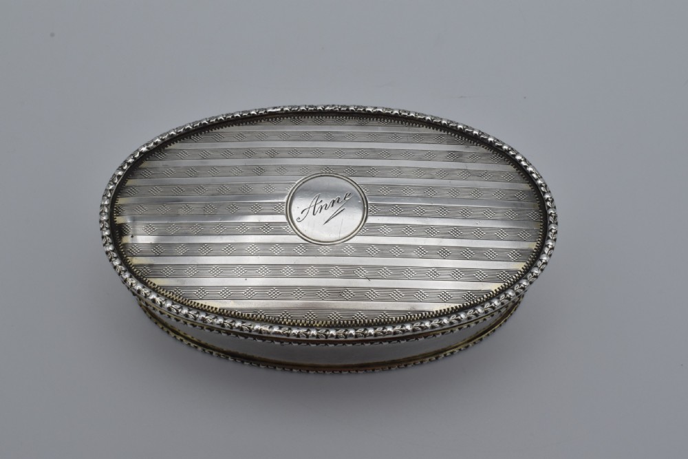 An oval antique sterling silver trinket box featuring engine-turned striped detailing on the lid, a central circular cartouche engraved with text, and beaded rims. View II