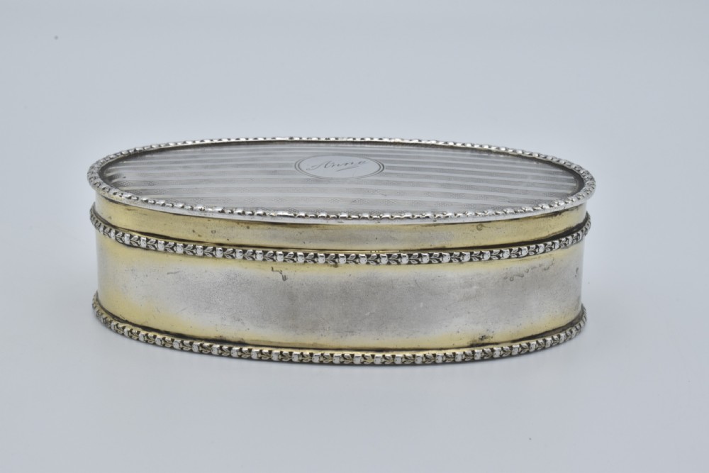 An oval antique sterling silver trinket box featuring engine-turned striped detailing on the lid, a central circular cartouche engraved with text, and beaded rims. View III