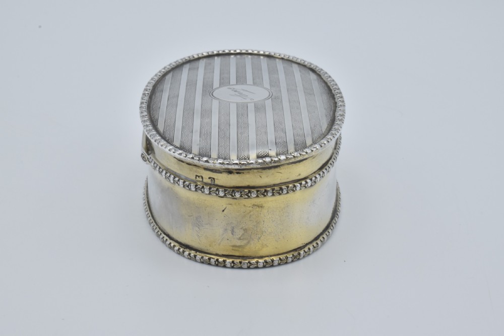 An oval antique sterling silver trinket box featuring engine-turned striped detailing on the lid, a central circular cartouche engraved with text, and beaded rims. View IV