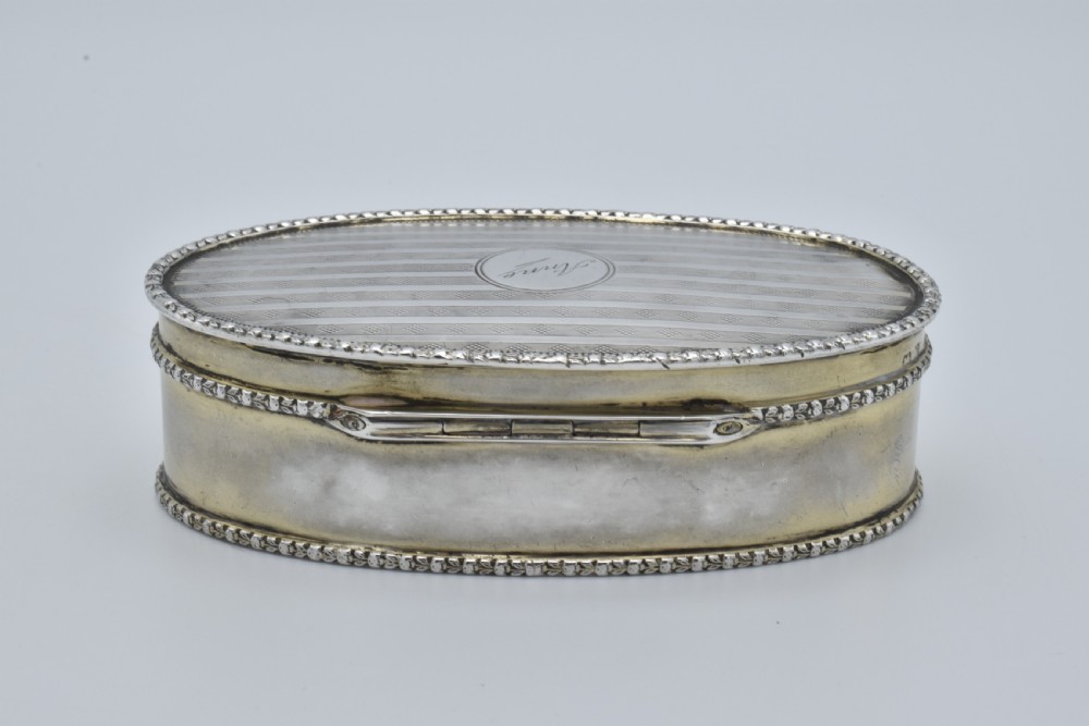 An oval antique sterling silver trinket box featuring engine-turned striped detailing on the lid, a central circular cartouche engraved with text, and beaded rims. View V