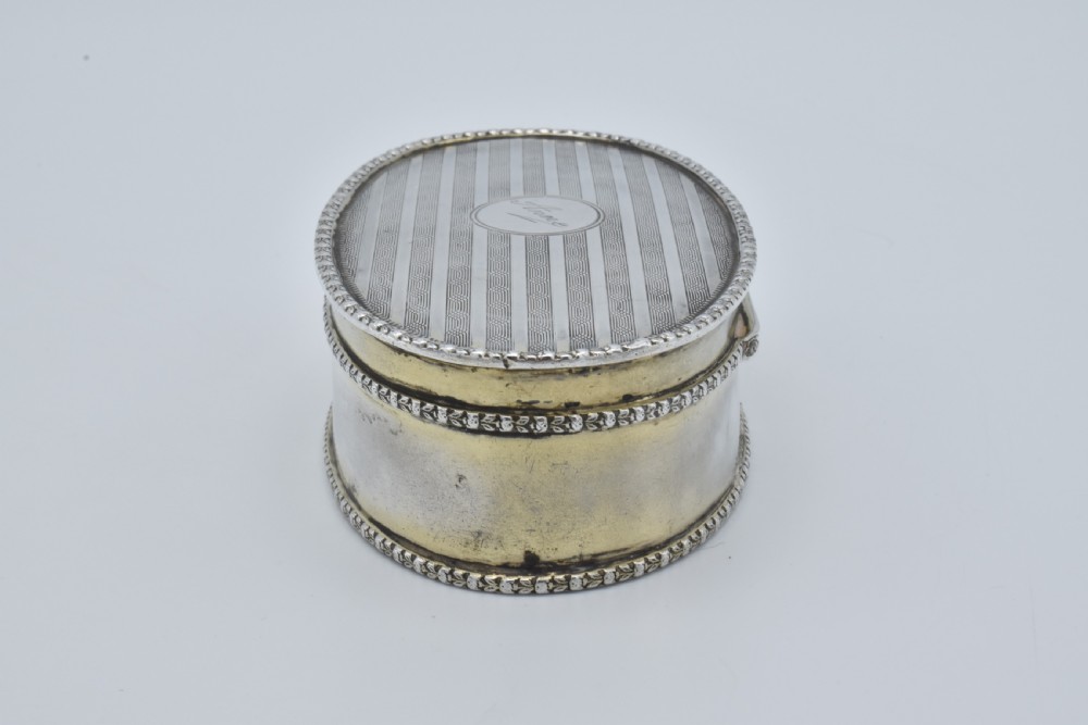 An oval antique sterling silver trinket box featuring engine-turned striped detailing on the lid, a central circular cartouche engraved with text, and beaded rims. View VI