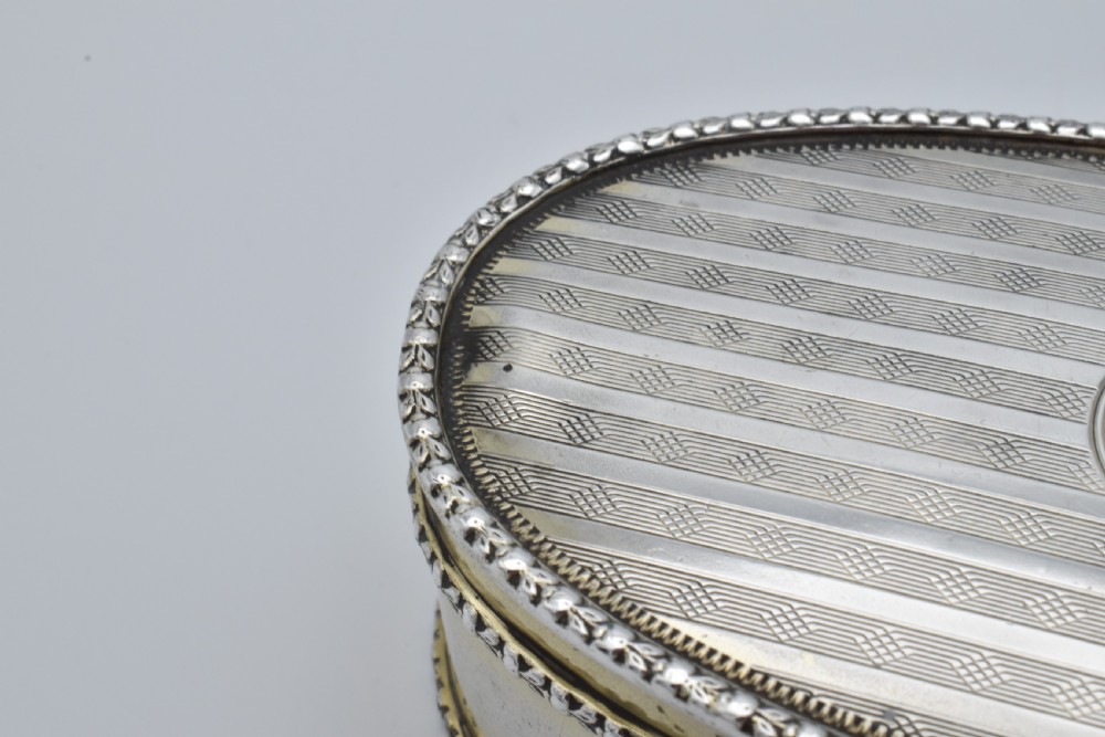 An oval antique sterling silver trinket box featuring engine-turned striped detailing on the lid, a central circular cartouche engraved with text, and beaded rims. View IX