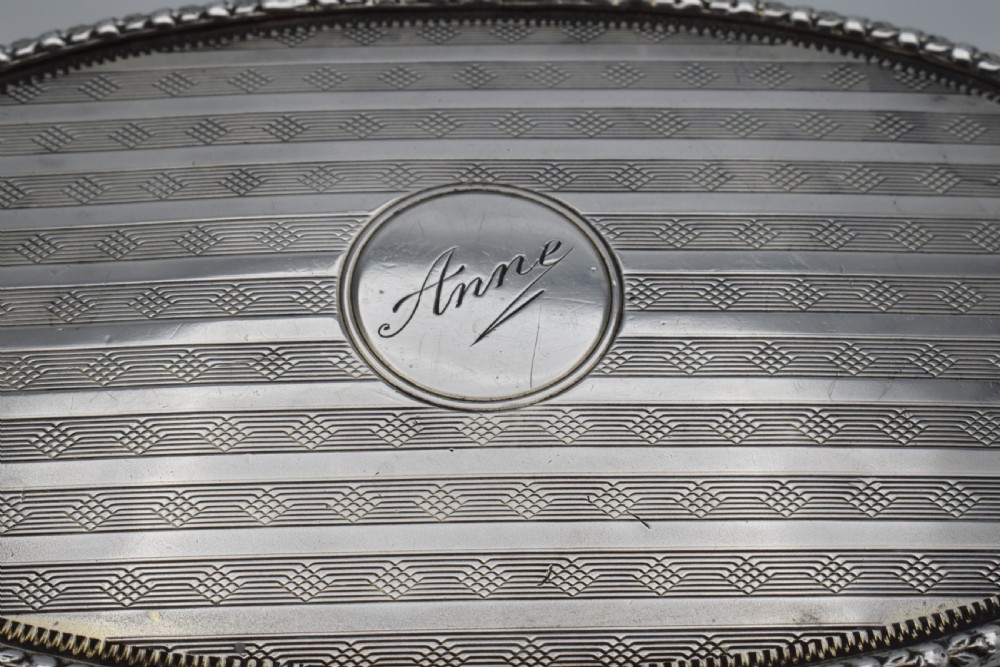An oval antique sterling silver trinket box featuring engine-turned striped detailing on the lid, a central circular cartouche engraved with text, and beaded rims. View X