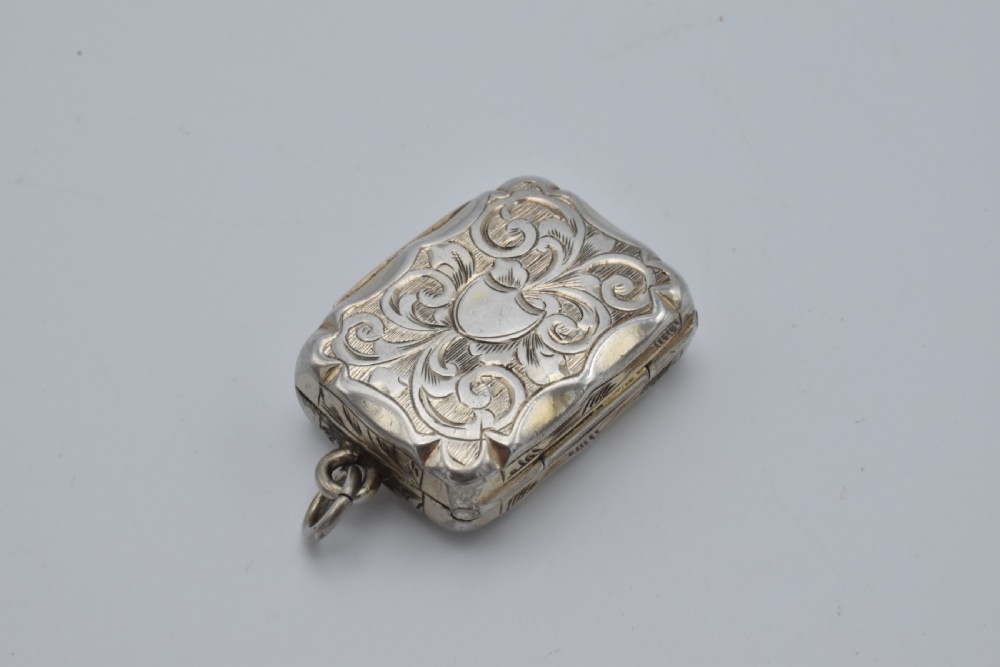 A small rectangular Victorian sterling silver vinaigrette featuring foliate engraving, a central shield cartouche, and a top suspension ring.