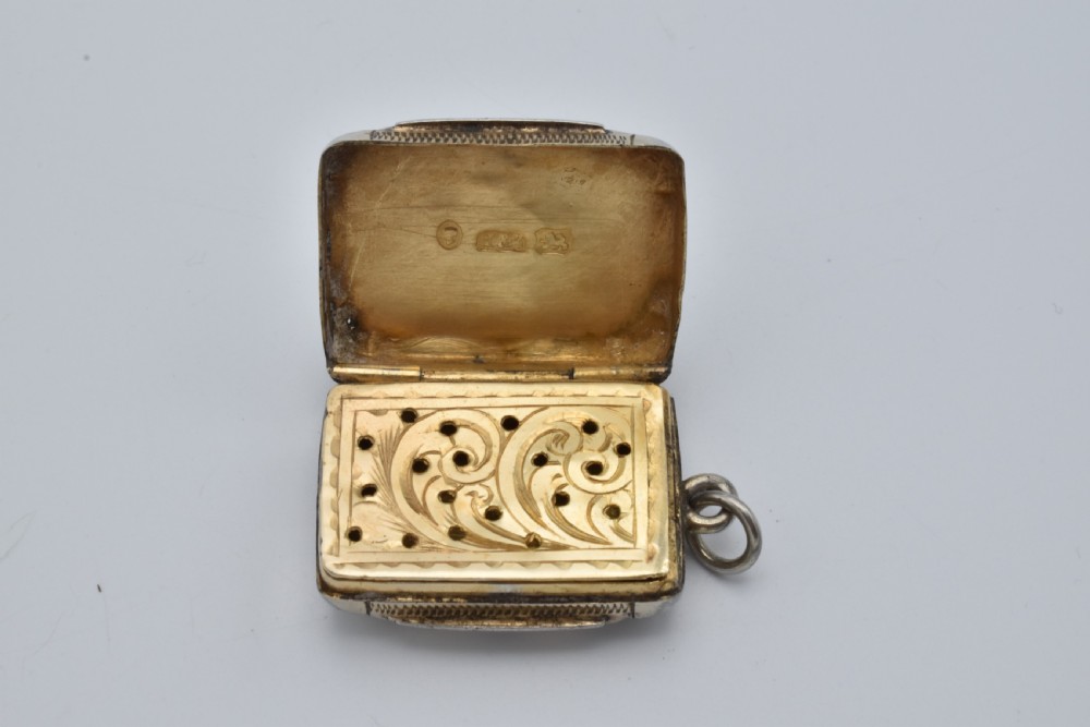 A small rectangular Victorian sterling silver vinaigrette featuring foliate engraving, a central shield cartouche, and a top suspension ring. View V