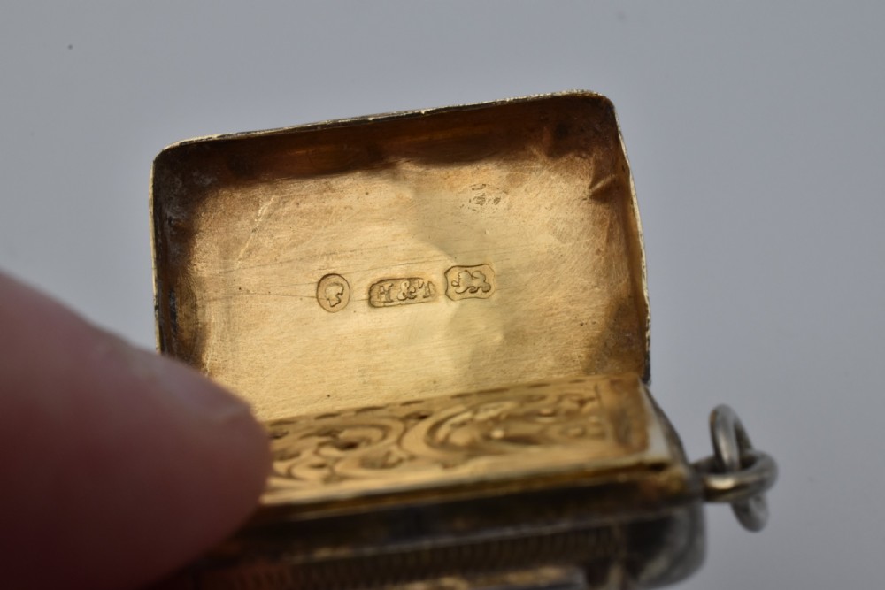 A small rectangular Victorian sterling silver vinaigrette featuring foliate engraving, a central shield cartouche, and a top suspension ring. View VI