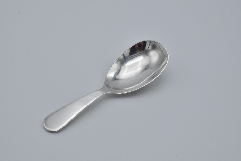 A plain sterling silver caddy spoon with a wide oval bowl and a smooth, unadorned handle resting on a neutral grey background.