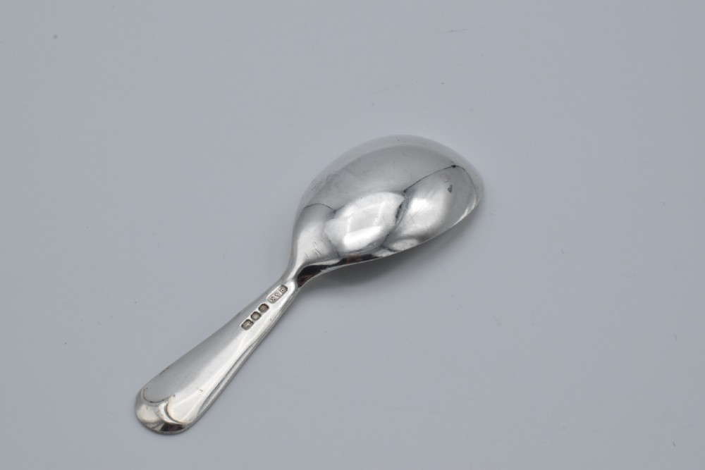 A plain sterling silver caddy spoon with a wide oval bowl and a smooth, unadorned handle resting on a neutral grey background. View II